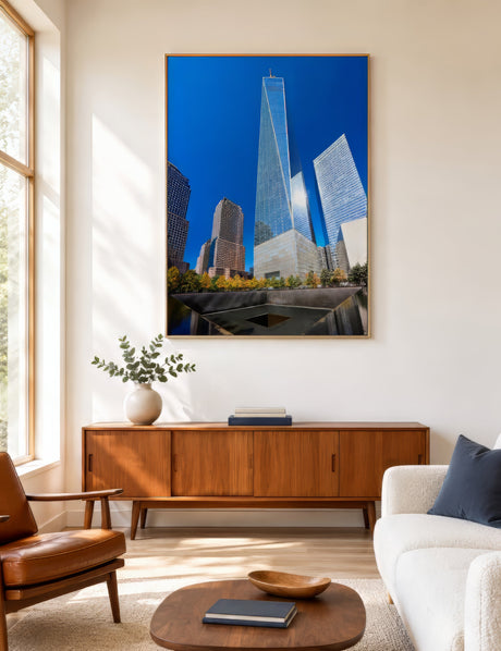 Photographic print of One World Trade Center and Manhattan skyline in blue and silver, framed beside a window in a bright small living room.