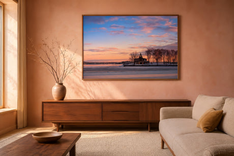 Romantic winter landscape print of a chapel by a frozen lake at sunset, walnut frame, above linen sofa in cozy golden-hour living room.