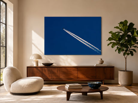 Modern deep blue canvas showing airplane contrail line, hung over walnut sideboard in airy contemporary living room with boucle chair