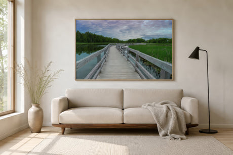 Minimalist landscape print of a boardwalk over marshland in muted greens and grays, slim white frame above a greige sofa in a bright Scandinavian living room.