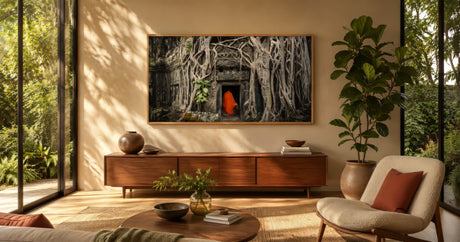 Framed canvas of a banyan-covered temple ruin with a saffron-robed monk, earthy greens and orange, in a sunlit sunroom with rattan seating.