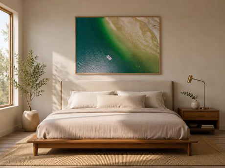 Minimalist aerial seascape canvas in turquoise green and sandy neutrals, framed above an upholstered bed in a calm morning-lit bedroom.