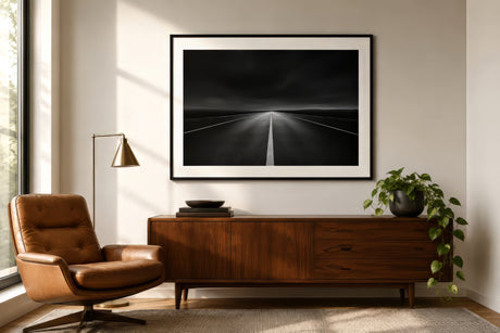 Black and white photograph of an open road disappearing into darkness, slim black frame with white mat, above a walnut console in a modern living room.