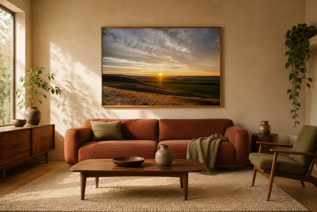Framed sunset landscape painting in ochre, terracotta and amber tones above a rust sofa in a warm earthy living room.