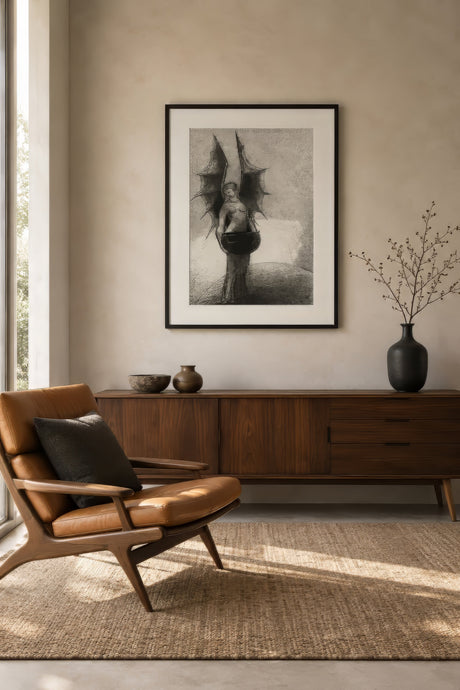 Dark gothic fallen angel print in charcoal and black, framed above a leather chair in a warmly lit study.