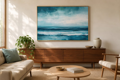 Blue coastal seascape painting with ocean horizon and soft waves, framed above a neutral sofa in a light-filled living room.