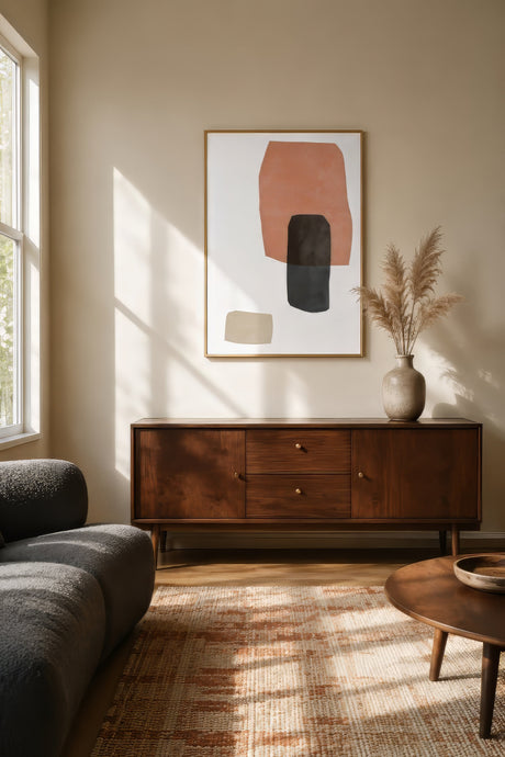 Abstract terracotta and black shapes on ivory in a slim walnut frame above a teak credenza in a sunlit mid-century living room.