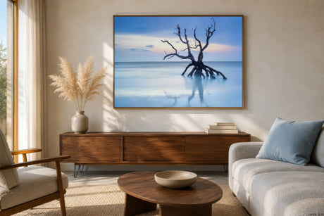 Coastal canvas of a bare driftwood tree on calm blue water, framed in wood above a linen sofa in an airy living room.