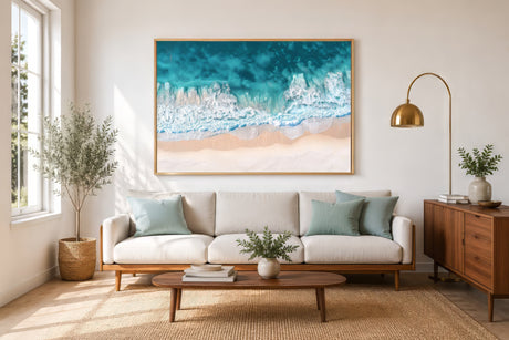 Aerial turquoise ocean and sandy beach canvas in wood frame above neutral sofa in calm coastal living room.
