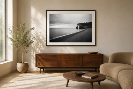 Black-and-white coastal sunset landscape photography canvas with cliff and shoreline, framed above a sofa in a calm modern living room.