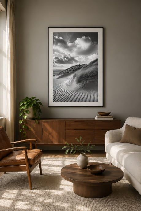 Black and white photography print of rippled sand dunes and clouds, framed in black above a walnut console in a modern living room.