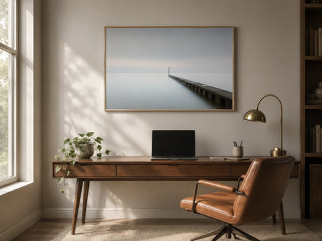 Minimalist horizon canvas of a pier over calm blue water, framed in wood above a mid-century desk in a sunlit office.