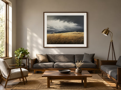 Vintage landscape print of a golden prairie under stormy gray clouds in a walnut frame above a sofa in a rustic living room.