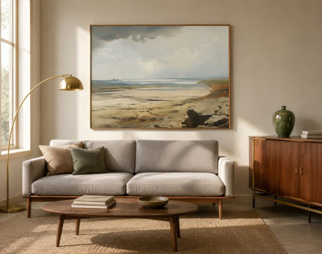 Vintage landscape painting of a coastal plain in beige and blue, framed above a neutral sofa in a warm modern living room.