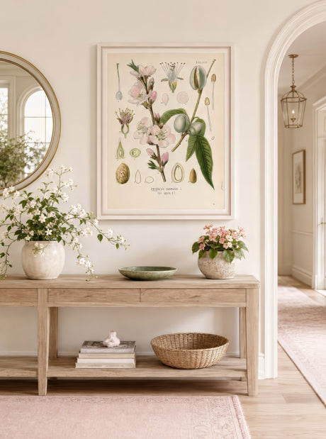 Botanical line art print of pink blossoms and green leaves framed above a light oak console with monstera and trailing plants in an airy entryway.