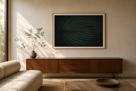 Black and white monstera leaf print with deep green tones and generous negative space, framed in light wood above a walnut console in a warm minimalist living room.