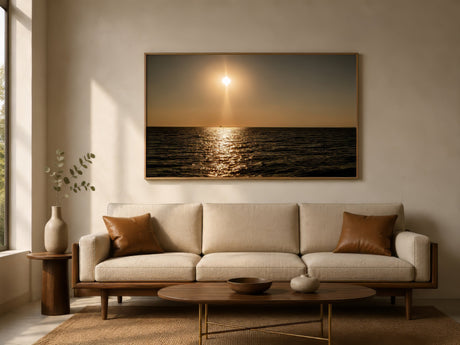Wide minimalist ocean sunset print in golden and bronze tones, framed above a neutral sofa in a calm modern living room.