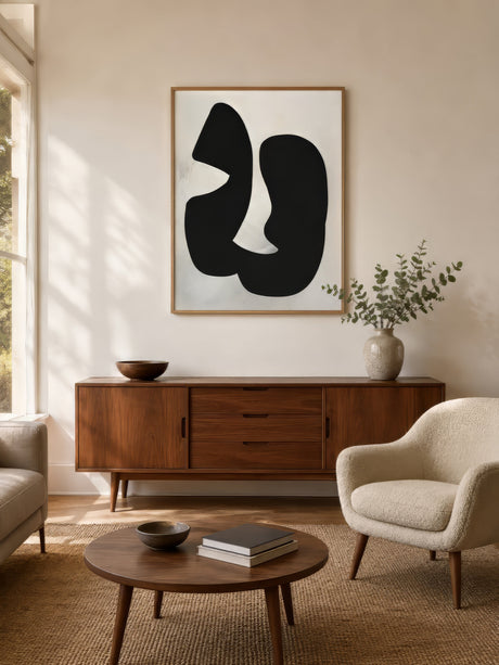 Black and white abstract organic-shape canvas in light wood frame, centered above a walnut sideboard in a calm modern living room.