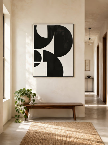 Black and ivory geometric abstract print with circular forms, framed in black, above a wooden bench in a warm minimalist hallway.