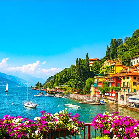 Colorful Lake Como waterfront village with sailboats on vibrant blue water, Italian hillside homes, and blooming flowers – premium wall art for home or office décor.