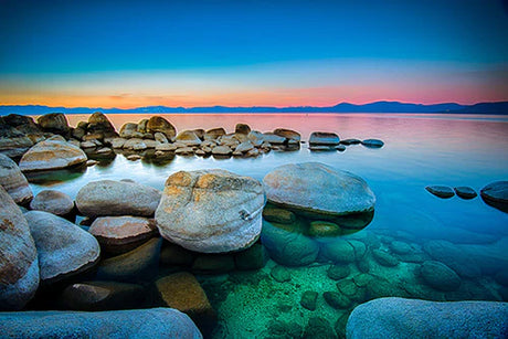 Dusk sunrise over Lake Tahoe displaying blue waters, boulders, and natural landscape reflection