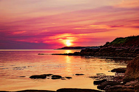 Wall art featuring a stunning sunset over Prince Edward Island's rocky coastline, with vibrant orange afterglow on the horizon.