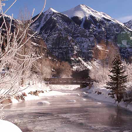 Snow-covered Telluride mountains with frozen river and rustic bridge in winter sunlight – serene Colorado landscape wall art for cozy home or lodge décor.