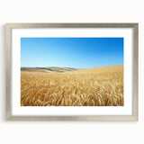 Framed landscape artwork showcasing a serene countryside scene, featuring lush green fields and a clear blue sky, with a silver mat providing an elegant touch.