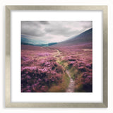 Framed landscape wall art depicting a serene countryside scene, featuring vivid greens and soft blues, with a silver mat and elegant frame, perfect for home decor.