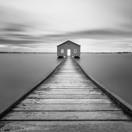 Fine art black and white photograph of a wooden dock leading to a boathouse over calm water