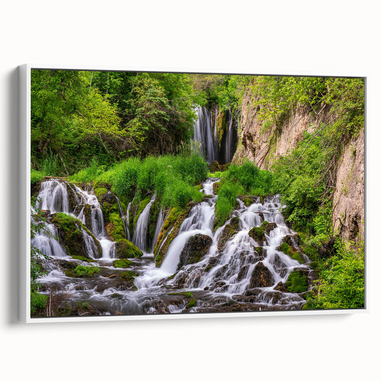 Framed canvas wall art showcasing a serene landscape of cascading waterfalls, surrounded by lush greenery, with a sleek white frame enhancing its elegance.