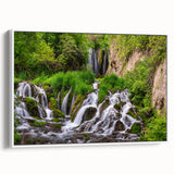 Framed canvas wall art showcasing a serene landscape of cascading waterfalls, surrounded by lush greenery, with a sleek white frame enhancing its elegance.
